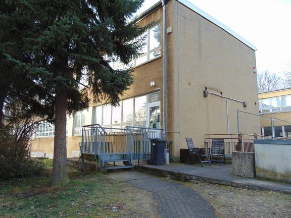 Auf dem Bild ist die Fassade des Jugendzimmers Legefeld zu sehen. Es sind der Eingang und ein großer Baum zu sehen,