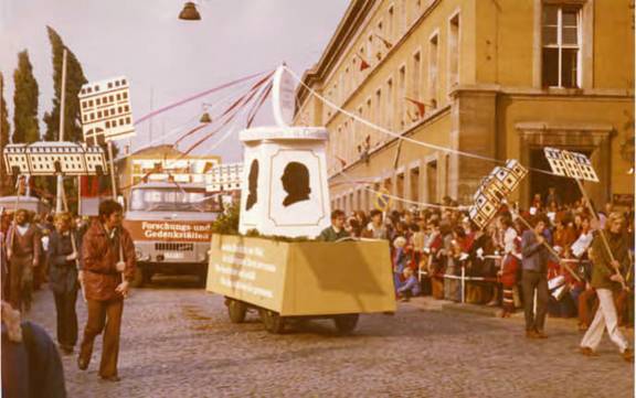 Festumzug „1000 Jahre Weimar“ am 12. Oktober 1975 mit Fahrzeugen der Nationalen Forschungs- und Gedenkstätten der klassischen deutschen Literatur. Flankierende Mitarbeiter tragen stilisierte Gedenkstätten-Darstellungen; hinter der Absperrung drängen sich Zuschauer. Aufnahme am Karl-Marx-Platz (heute Jorge-Semprún-Platz).