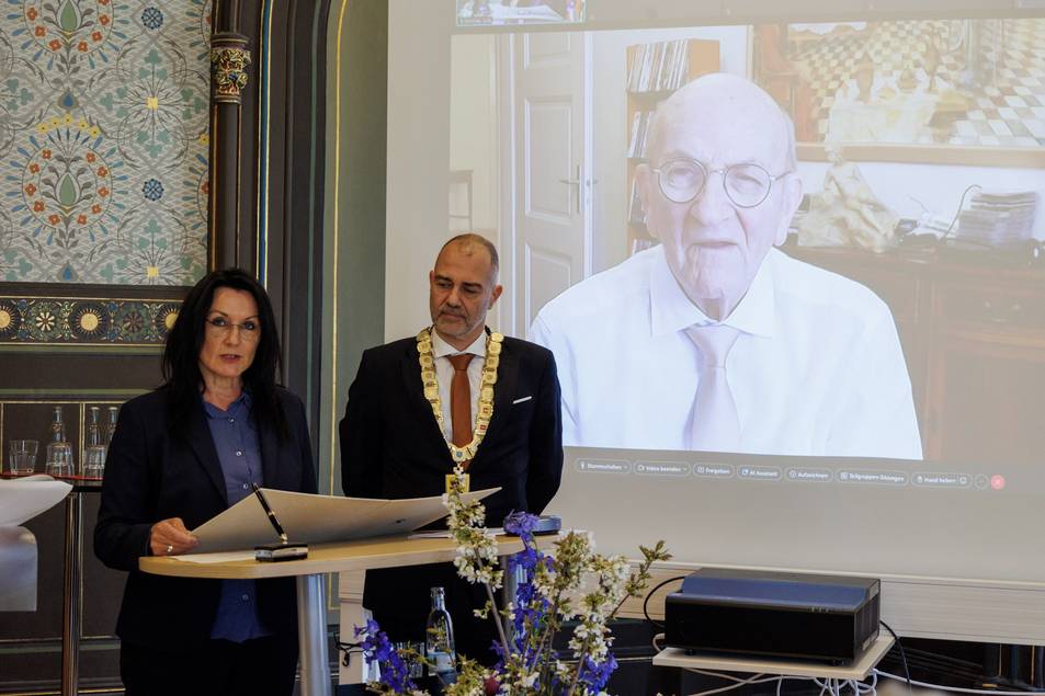 Katrin Götz und Oberbürgermeister Peter Kleine beim Verlesen eines Urkundentextes, im Hintergrund Isaak Goldblum via Videokonferenz zugeschaltet.