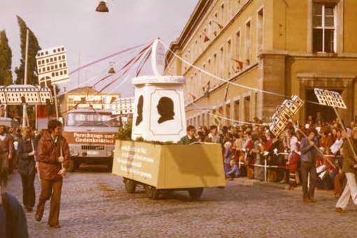 Festumzug „1000 Jahre Weimar“ am 12. Oktober 1975 mit Fahrzeugen der Nationalen Forschungs- und Gedenkstätten der klassischen deutschen Literatur. Flankierende Mitarbeiter tragen stilisierte Gedenkstätten-Darstellungen; hinter der Absperrung drängen sich Zuschauer. Aufnahme am Karl-Marx-Platz (heute Jorge-Semprún-Platz).