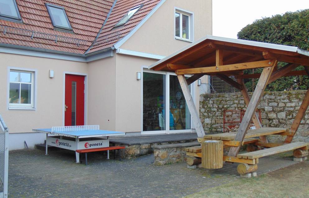 Auf dem Bild ist die Terasse mit Sitzgurppe und die rote Eingangstür des Jugendzimmers Süßenborn zu sehen.